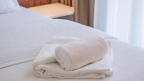A neatly made bed with a folded white towel, a bedside table with a lamp, and a window with curtains. - Hotel Onogošt (Nikšić)