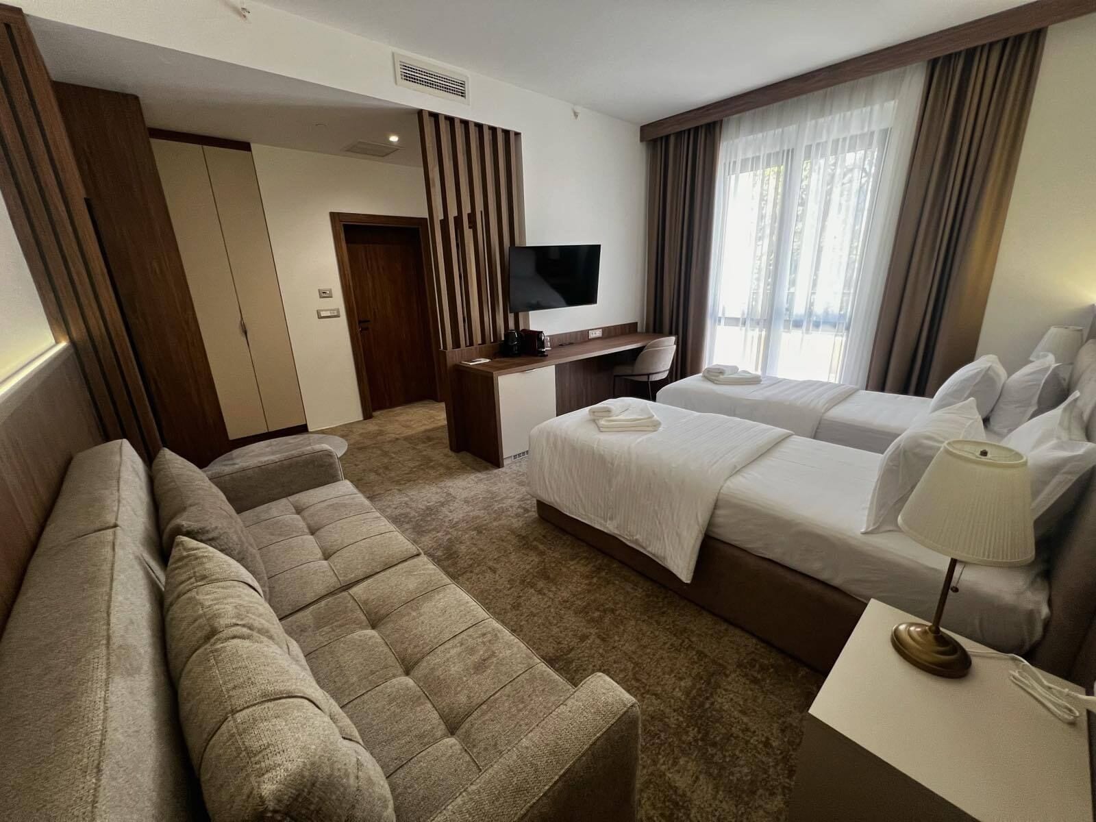 Une chambre d’hôtel avec un canapé, deux lits, un bureau avec une télévision et une fenêtre avec des rideaux.