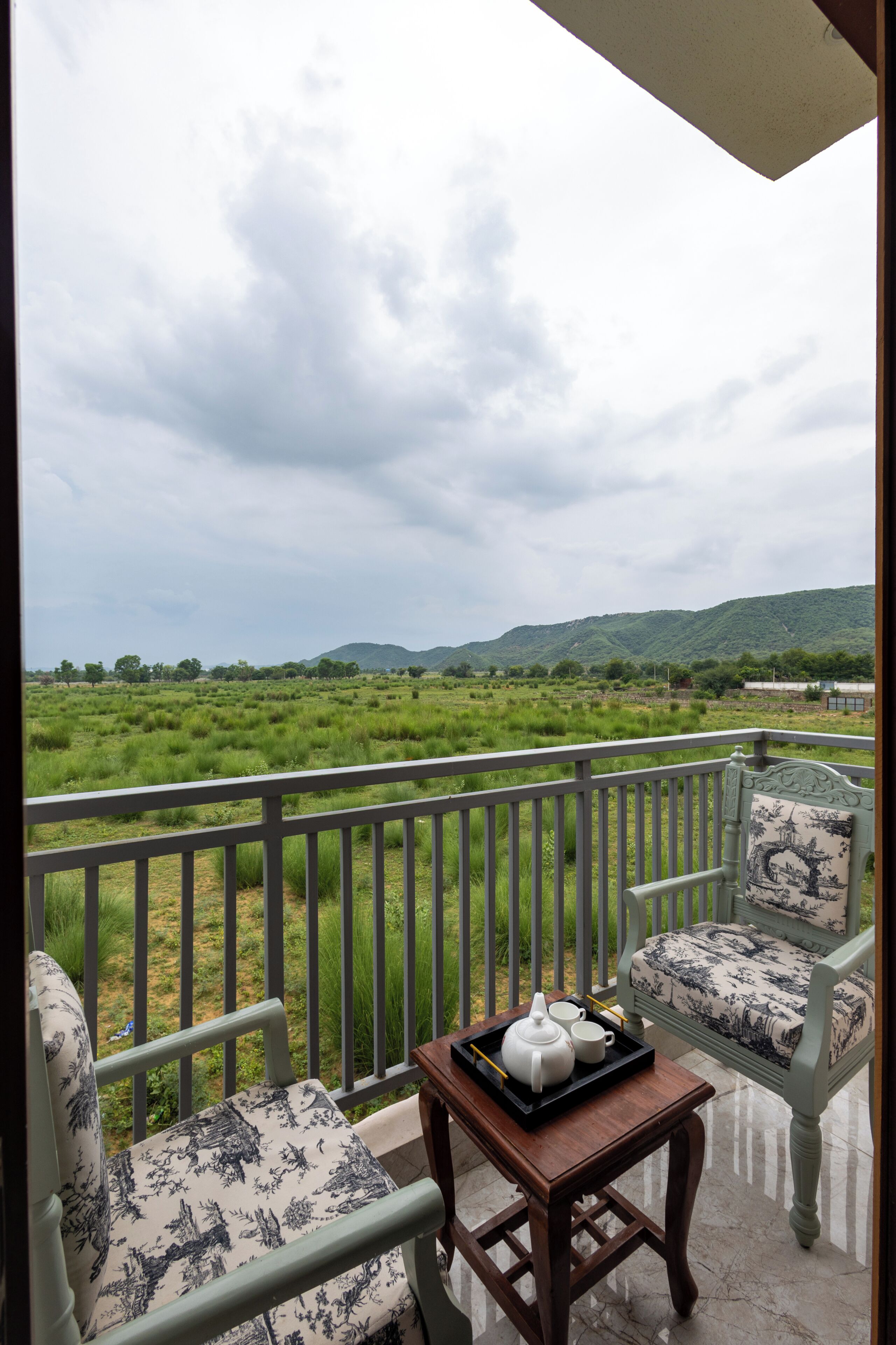 A balcony with a view of a green landscape and hills, a table with a teapot and cups, and a chair with a patterned cushion.