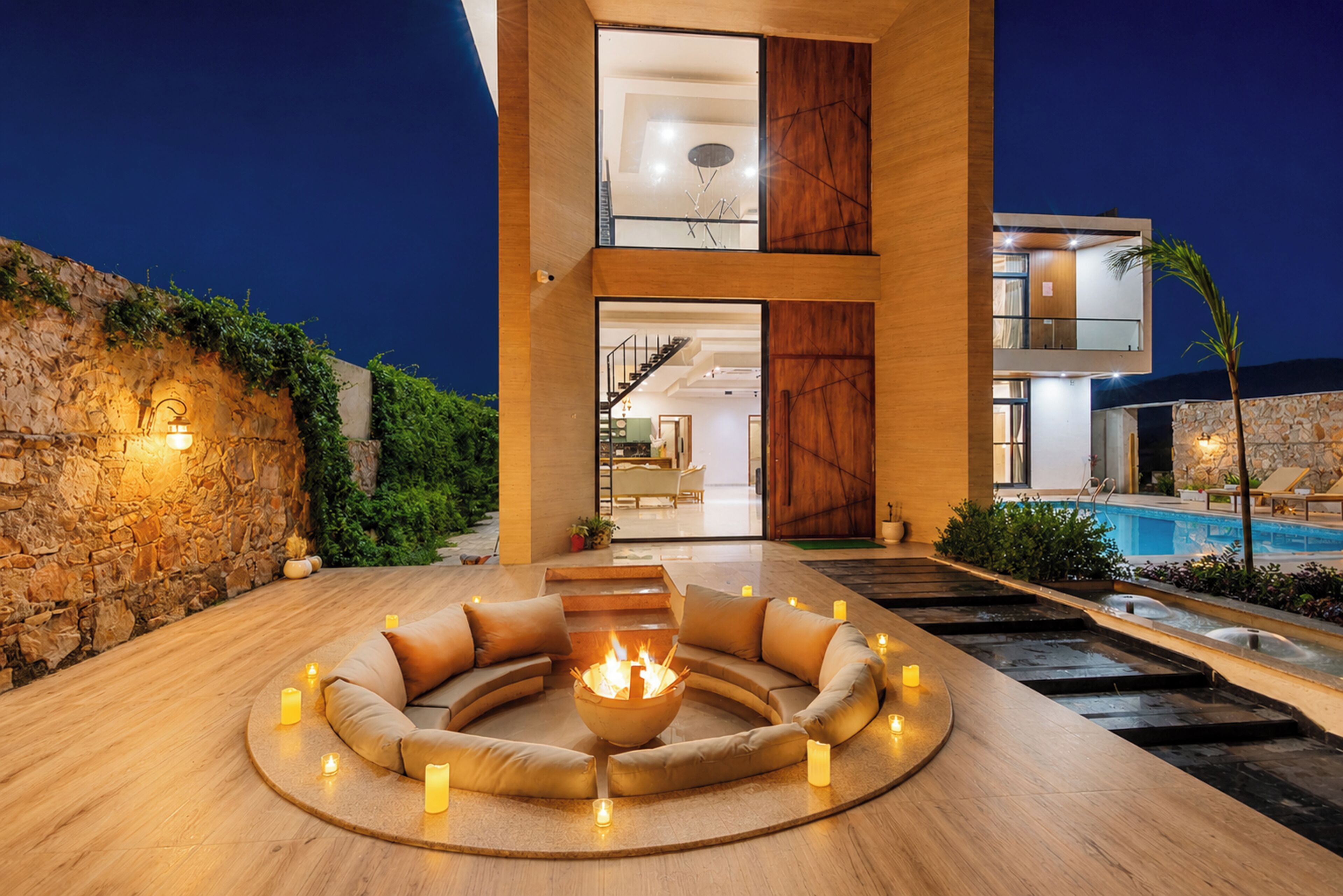 Un espacio exterior moderno con área de estar y una chimenea, rodeado de velas, frente a un edificio contemporáneo con piscina.
