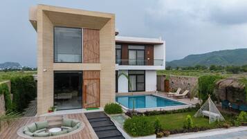 A modern two-story house with a swimming pool, outdoor seating, and a view of the surrounding landscape.