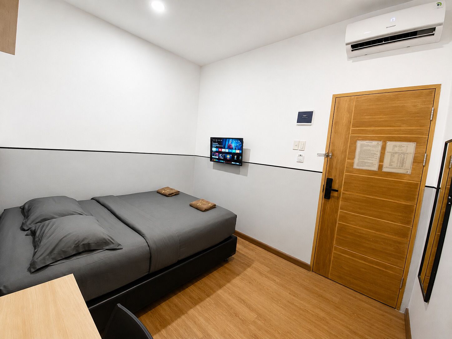 A small, modern room with a single bed, a mounted TV, an air conditioning unit, and a wooden door with a notice.
