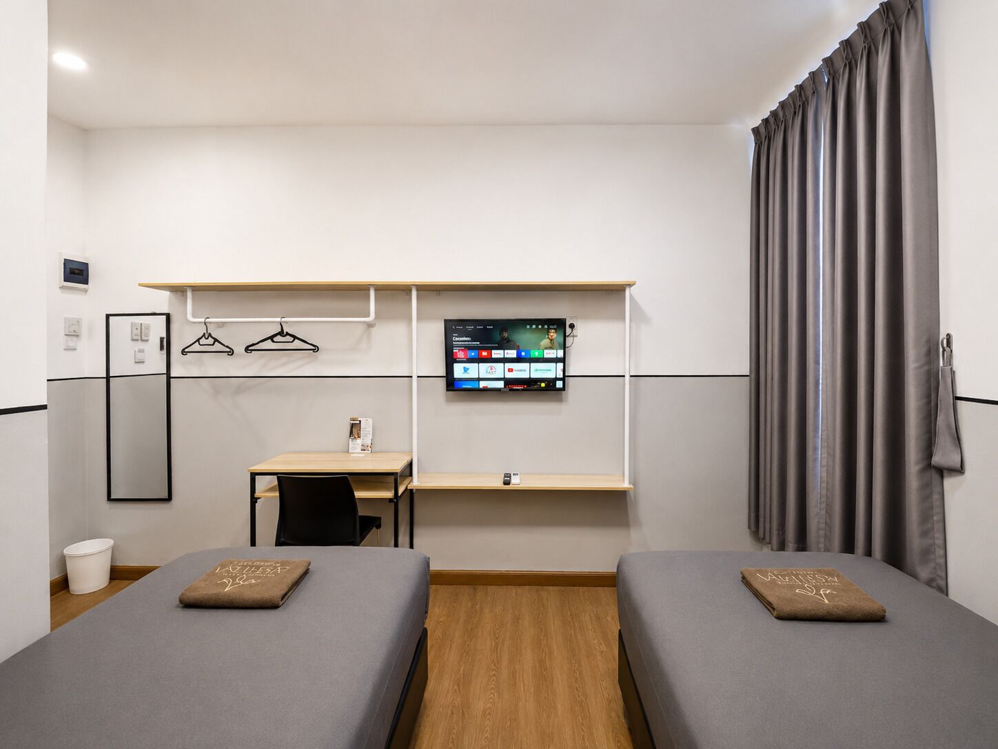 Een hotelkamer met twee enkele bedden, een bureau, een stoel, een televisie en een groot raam met gordijnen.