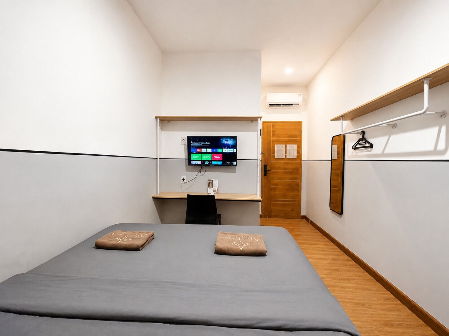 Une chambre d’hôtel avec un lit, un bureau doté d’un écran d’ordinateur, une porte en bois et un climatiseur fixé au mur.