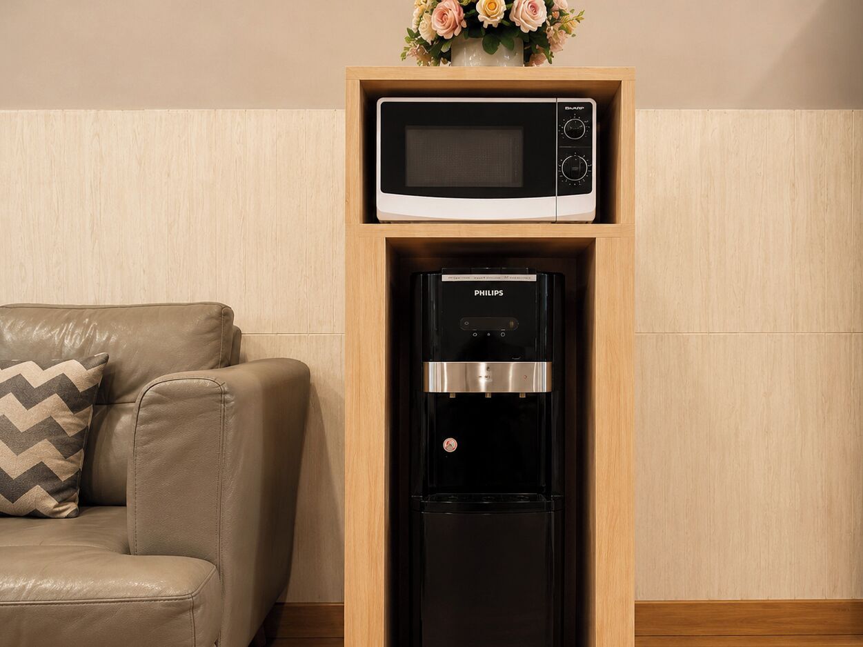 A compact kitchen setup with a microwave and a water dispenser in a wooden cabinet, a leather sofa with a patterned pillow, and a vase with flowers on the wall.