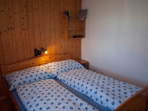A wooden room with a bed, a shelf with books, and a wall-mounted lamp.