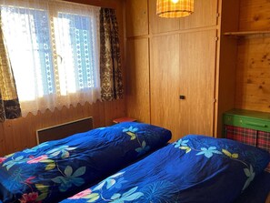 A room with two beds, wooden walls, a window with curtains, and a small table. - Chalet Bambi Appartement/Fewo, Dusche, WC 4-Bettwohnung by Interhome (Rosswald)