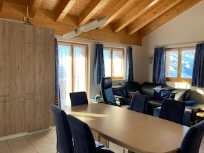 Ein Zimmer mit Holzbalkendecke, einem Esstisch mit Stühlen, einem Sofa und Blick auf Berge durch die Fenster.