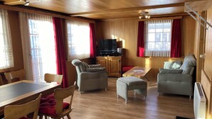 Una habitación con paneles de madera, una mesa de comedor, dos sillones, un sofá, un televisor y un radiador.