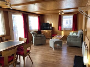 Ein Zimmer mit Holzvertäfelung, einem Esstisch, zwei Sesseln, einem Sofa, einem Fernseher und einem Heizkörper.