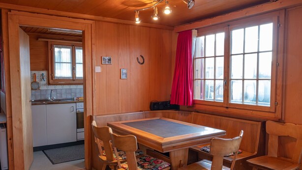 Un interior de madera con una mesa de comedor y sillas, una zona de cocina y un ventanal con cortinas.
