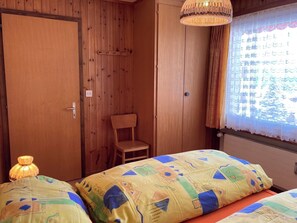 A room with wooden walls, a single bed with patterned bedding, a chair, a hanging light fixture, and a window with curtains. - Chalet Sunnuterrasse Appartement/Fewo, Dusche, WC 4-Zimmerwohnung Dusche, WC by Interhome (Rosswald)
