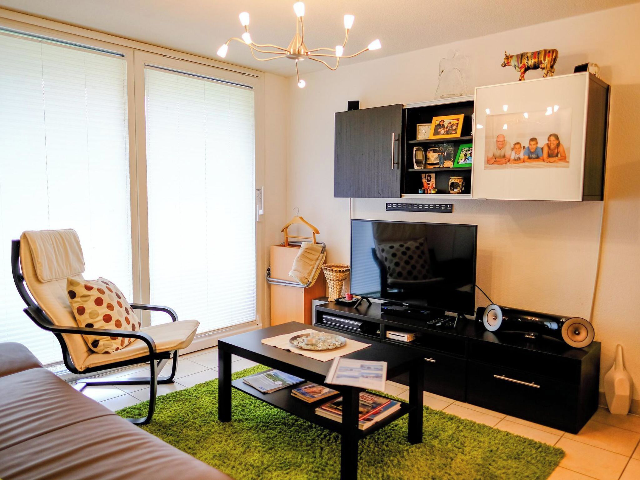 Een moderne woonkamer met een bank, een fauteuil, een salontafel, een televisie, een kast met foto's en een decoratieve plafondlamp.