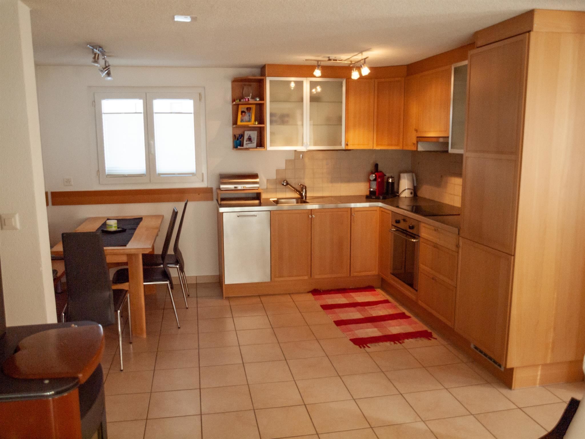 A compact kitchen with wooden cabinets, a dining table with chairs, and a red and white checkered rug.