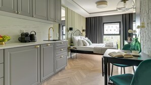 Moderni hotellihuone, jossa on pieni keittiönurkkaus, valkoisilla lakanoilla varustettu sänky, ruokailutila pöytineen ja tuoleineen sekä näkymä ulos.