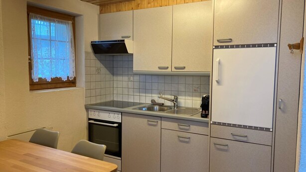 A compact kitchen with a wooden ceiling, a window with lace curtains, a built-in oven, a sink, and a white refrigerator. - Chalet Gerbera Appartement/Fewo, Bad, WC 5-Bettwohnung by Interhome (Rosswald)