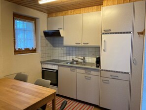 Una cocina compacta con techo de madera, una ventana con cortinas de encaje, horno empotrado, pileta y un refrigerador blanco.
