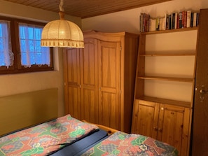 A bedroom with a bed, a wooden wardrobe, a shelf with books, and a window with curtains. - Chalet Gerbera Appartement/Fewo, Bad, WC 5-Bettwohnung by Interhome (Rosswald)