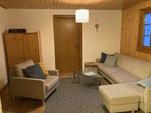 Una sala de estar con sofá, sillón, mesa de centro de vidrio y aparador de madera.