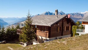 Eine Holzhütte mit geneigtem Dach, Schornstein und Blick auf schneebedeckte Berge im Hintergrund.