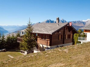 Eine Holzhütte mit geneigtem Dach, Schornstein und Blick auf schneebedeckte Berge im Hintergrund.