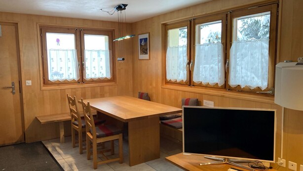 Ein Zimmer mit einem Holzespeisetisch, Stühlen, einem Fernseher und Fenstern mit Vorhängen.
