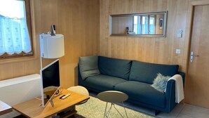 Ein Zimmer mit Holzboden, einem grünen Sofa, einem kleinen runden Tisch, einer Lampe und einem Fenster mit Vorhängen.