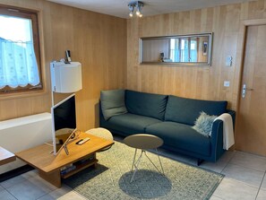Ein Zimmer mit Holzboden, einem grünen Sofa, einem kleinen runden Tisch, einer Lampe und einem Fenster mit Vorhängen.