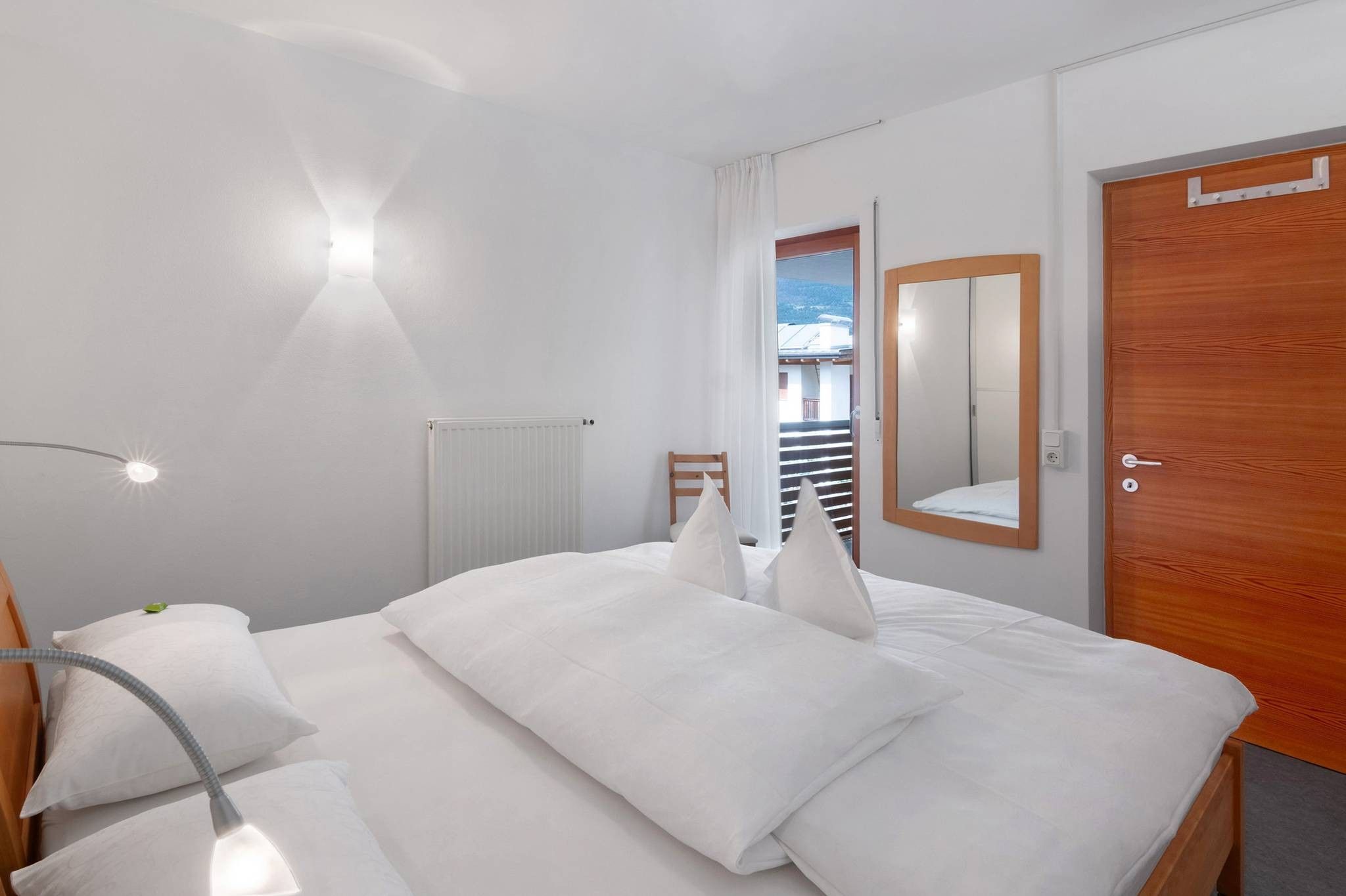 Ein Hotelzimmer mit einem Bett, einem Sessel, einem Schreibtisch, einer Lampe, einem Heizkörper, einem Spiegel und einer Tür.
