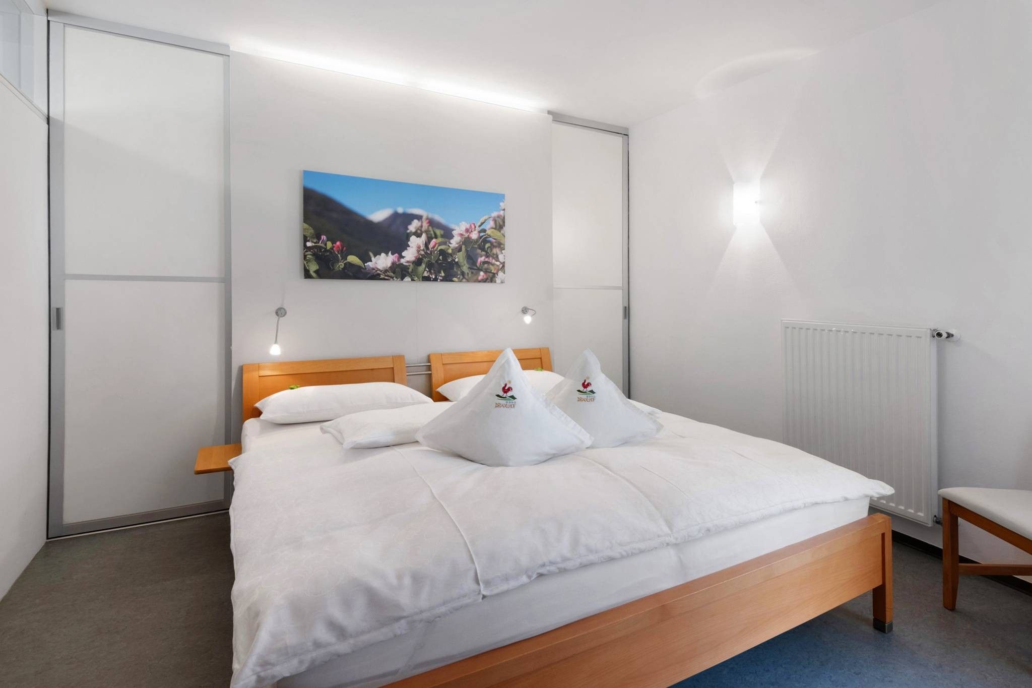 Ein Schlafzimmer mit einem Bett, Kissen, einem Holzkopfende, einem an der Wand hängenden Bild, einem Nachttisch und einem Heizkörper.