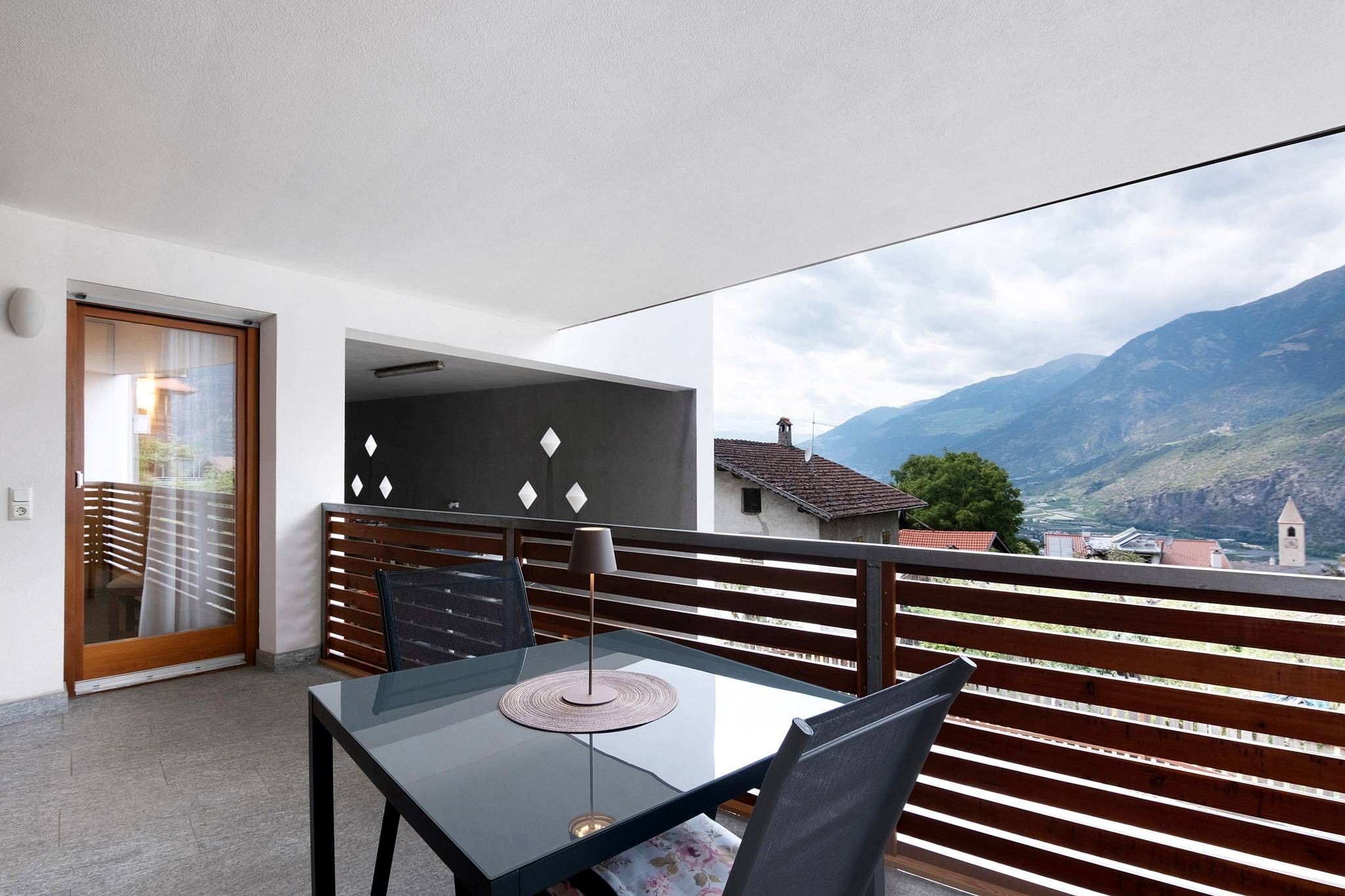 A balcony with a table and chairs, a view of mountains, and a building with a red roof.