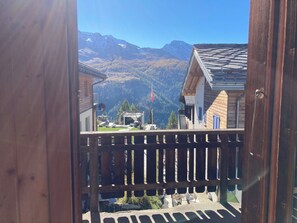 A wooden balcony with a view of a mountain landscape, a flag, and a building with a sloped roof.