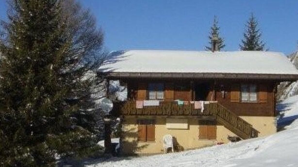 Eine Holzhütte mit schneebedecktem Dach, umgeben von Bäumen und einem verschneiten Weg, der zu ihr führt.