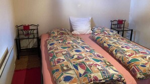 Ein kleines Zimmer mit zwei Einzelbetten, einem roten Teppich, einem Tisch mit einem roten Stuhl und einem gerahmten Bild an der Wand.