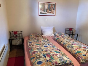 Ein kleines Zimmer mit zwei Einzelbetten, einem roten Teppich, einem Tisch mit einem roten Stuhl und einem gerahmten Bild an der Wand.
