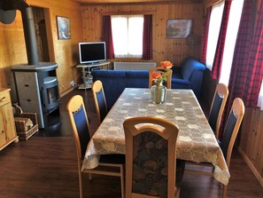 A cozy cabin interior with a dining table, chairs, a sofa, a television, and a wood-burning stove.