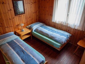 Ein Zimmer mit zwei Einzelbetten, Holzwänden, einem Nachttisch mit Lampe und einem Fenster mit durchscheinenden Vorhängen.
