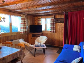 Ein gemütliches Blockhaushaus-Interieur mit Holzbalkendecke, einem Fernseher, einem Esstisch und einer Couch.