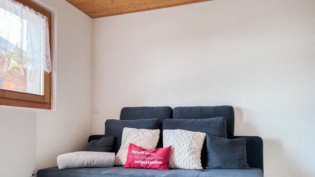 Ein dunkelblaues Sofa mit vielen Kissen, ein rotes Kopfkissen mit Schriftzug und eine Stern-Dekoration am Fenster.