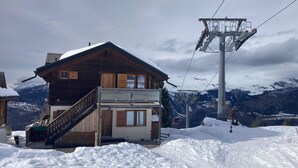 Eine Holzchalet mit Balkon, dahinter eine Seilbahn und schneebedeckte Berge.