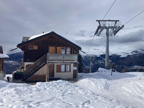 Eine Holzchalet mit Balkon, dahinter eine Seilbahn und schneebedeckte Berge.