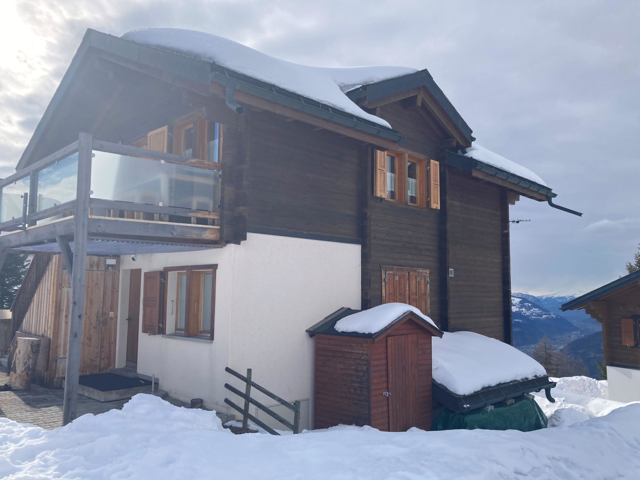 A two-story wooden chalet with a balcony, snow-covered roof, and a mountain backdrop.