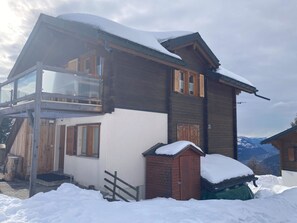 Eine zweistöckige Holzhütte mit Balkon, verschneitem Dach und Bergblick.