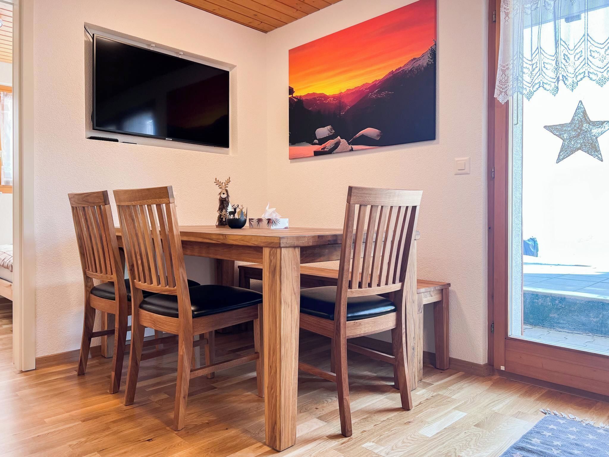 A dining area with a wooden table and chairs, a flat-screen TV mounted on the wall, and a painting of a sunset.