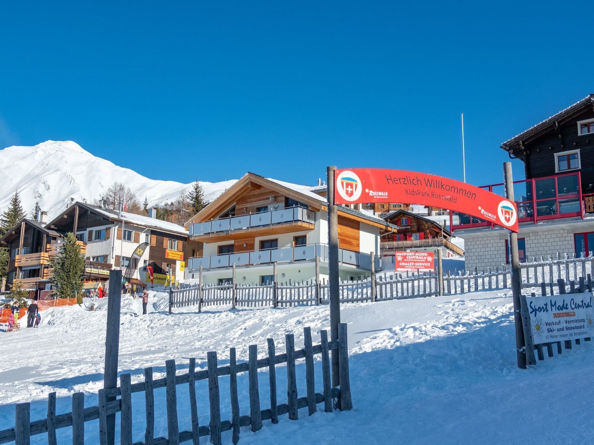 Een skigebied met een houten hek, een rood bord en gebouwen met sneeuw bedekte daken.