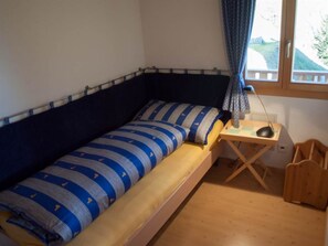 A small, single-bed room with a wooden floor, a desk with a chair, and a window with curtains. - Chalet Bergkristall 4-Bettwohnung, Dusche, WC 4-Bettwohnung, Dusche, WC by Interhome (Rosswald)