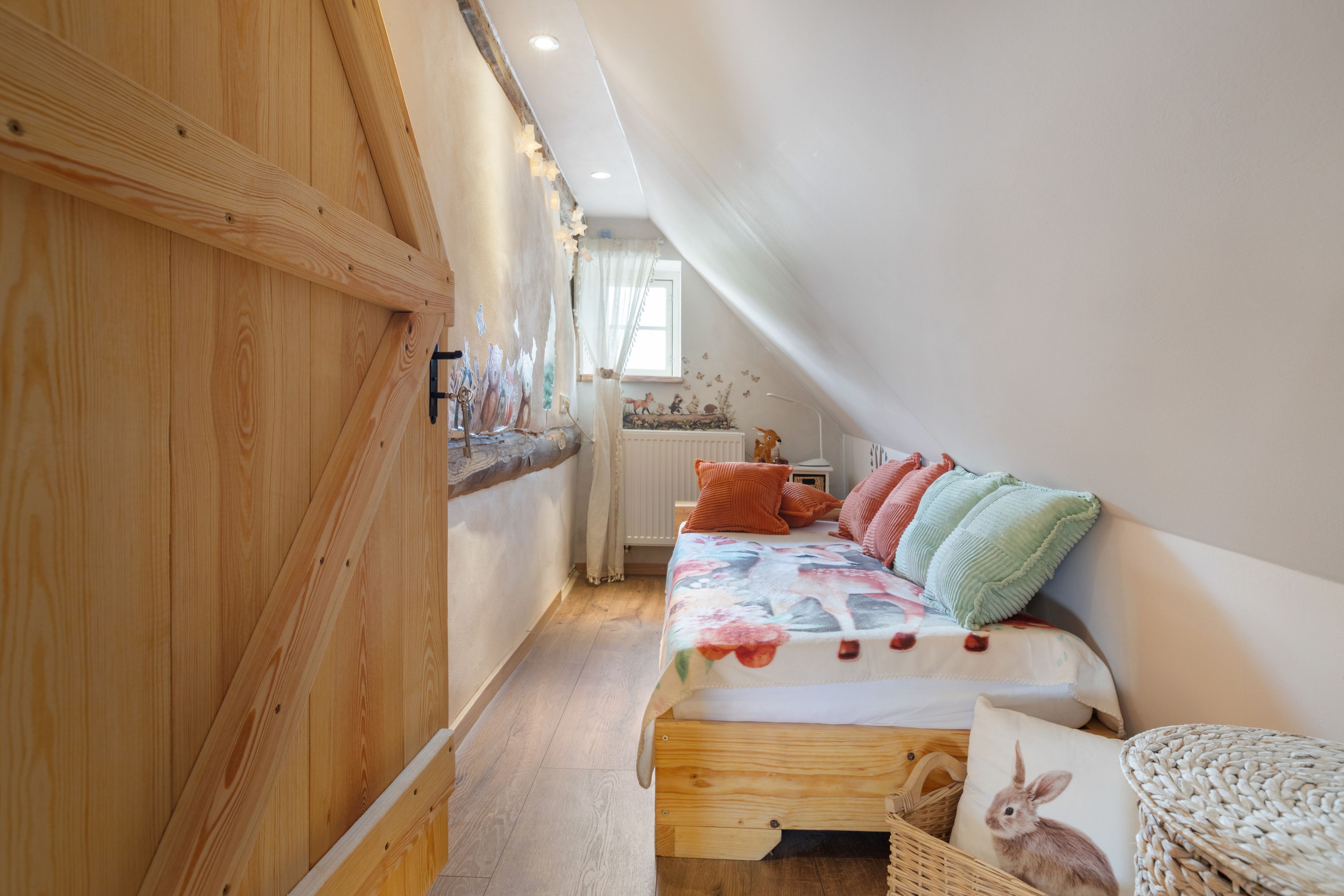 A narrow room with a sloped ceiling, wooden walls, a bed with colorful bedding, and a small window.