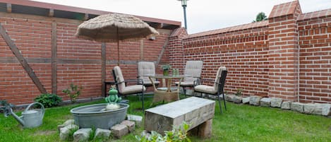 A backyard with a brick wall, a metal table, four chairs, a thatched umbrella, a metal watering can, and a frog statue.