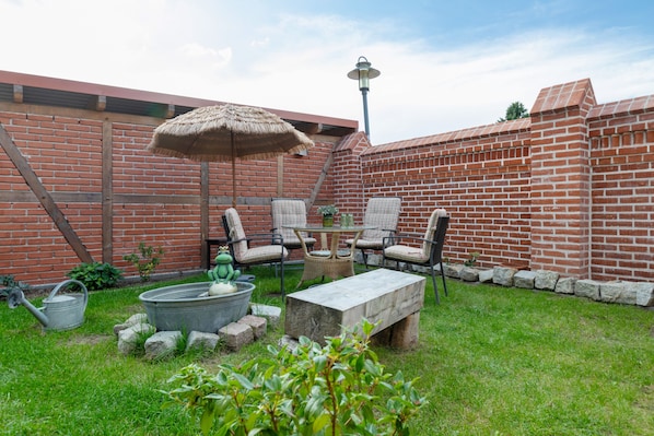 A backyard with a brick wall, a metal table, four chairs, a thatched umbrella, a metal watering can, and a frog statue.