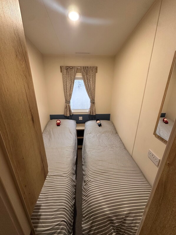 A small room with two single beds, a window with curtains, a mirror, and a light fixture on the ceiling.